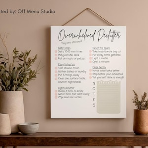 May include: A white canvas print with the words "Overwhelmed Declutter" and a list of decluttering tips. The print is hanging on a wall above a wooden table with decorative vases and plants.