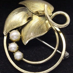 May include: A gold-toned brooch featuring three faux pearls and a leaf design. The brooch has a pin closure on the back and a decorative swirl element. The leaves have a textured finish.
