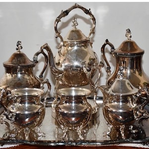 May include: A silver tea set, including a teapot, creamer, sugar bowl, and two teacups, is displayed on a silver tray. The set has ornate handles and lids, reflecting light. The tray has decorative edges, and the set is placed on a wooden surface.