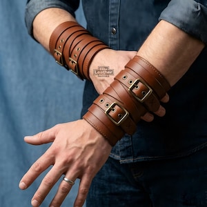 Könnte beinhalten: Ein Paar braune Leder-Armstulpen mit mehreren Riemen und Schnallen aus antikem messingfarbenem Metall. Jede Stulpe umschließt den Unterarm mit verstellbaren Ösenverschlüssen. Der Text Elite Leather Attire ist auf der Lederoberfläche aufgedruckt.
