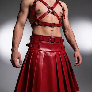 May include: A red leather kilt with pleats and a matching harness. The harness features straps and metal rings. The kilt has a belt with buckles. The outfit is a bold fashion statement.