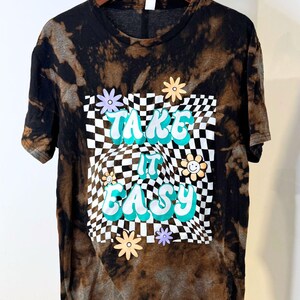 Könnte beinhalten: Schwarz-braunes Batik-T-Shirt mit den Worten "TAKE IT EASY" in Türkis und Weiß, vor einem karierten Hintergrund. Das Shirt ist mit Blumengrafiken verziert.