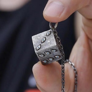 May include: A silver-colored pendant in the shape of a die, featuring eye-shaped designs in place of the traditional dots. The pendant is suspended from a chain. The die is held between fingers.