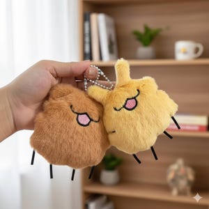 Pode incluir: Dois chaveiros de pelúcia, um castanho e outro amarelo, são exibidos. Cada chaveiro tem uma textura fofa, olhos bordados e uma boca rosa. Linhas pretas servem como pernas. Os chaveiros são presos a uma corrente prateada.