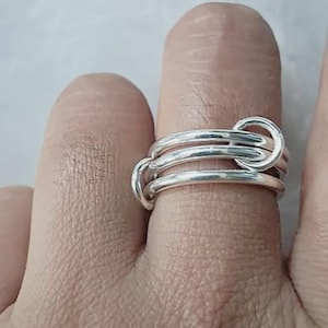 May include: A silver-colored ring featuring three interconnected, smooth, rounded bands that overlap and loop together. The metal has a polished, reflective finish, creating a continuous, layered appearance around the finger it is worn on.