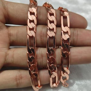 Pode incluir: Três pulseiras de corrente figaro em tom de ouro rosa, com um padrão repetitivo de três elos circulares pequenos seguidos por um elo oval alongado. As superfícies metálicas são polidas e refletoras, com um tom metálico quente de cobre.