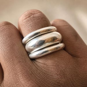 Puede incluir: Un conjunto de tres anillos de plata apilados con un acabado liso y pulido. Los anillos presentan bandas redondeadas y abovedadas de diferentes grosores, creando una apariencia de capas cuando se usan juntos en un mismo dedo.