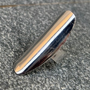 Pode incluir: Uma cobertura de unha prateada com ponta arredondada. A cobertura da unha é altamente reflexiva, mostrando o ambiente circundante. A cobertura da unha é montada em uma base retangular transparente.