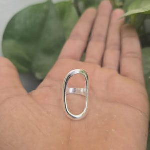 May include: A silver ring with an oval design. The ring is a solid piece of metal with a smooth, polished finish. The ring is shown on a hand with a green background.