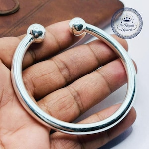 May include: A silver bangle bracelet with a rounded design and two ball-shaped ends. The bracelet is held in a hand, and the background is a leather surface. The Royal Spark logo is visible in the upper right corner.