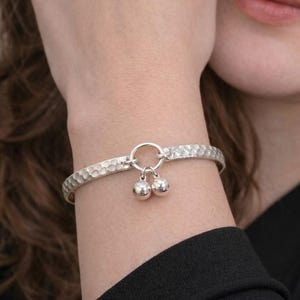 May include: A silver-toned bangle bracelet featuring a hammered, textured surface. The bracelet includes a central circular ring connector that holds two small, smooth, spherical charms that hang down from the main band of the jewelry piece.