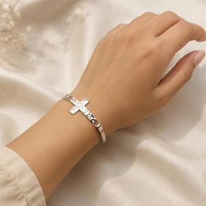 May include: A silver-toned metal bangle bracelet featuring a central cross charm with a hammered, textured finish. The bracelet has a slim, open-ended design that connects to the cross, creating a continuous circular shape around the wrist.