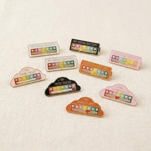 May include: A collection of enamel pins with the phrases "MY SOCIAL BATTERY" and "MOOD TRACKER". The pins feature a rainbow color scheme with a lightning bolt or a heart. The pins are in various shapes and colors, including white, black, pink, and orange.