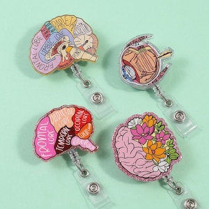 May include: Four medical-themed retractable badge holders. Designs include a brain diagram, an eye diagram, a brain diagram with labeled lobes, and a brain with floral accents. Each badge has a clear plastic strap and a silver-coloured clip.