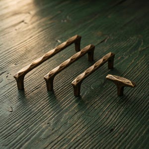 Può includere: Un set di quattro maniglie per mobili in metallo con finitura bronzo scuro, caratterizzate da una superficie martellata e testurizzata con un design ritorto e sfaccettato. La collezione include tre maniglie a barra di varie lunghezze e un pomello.