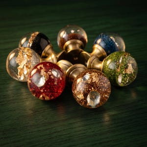 May include: A collection of spherical cabinet knobs featuring clear resin spheres filled with various colored glitters and metallic gold flakes. Each knob is attached to a circular brass-toned metal base, showcasing red, green, blue, black, and gold-toned designs.