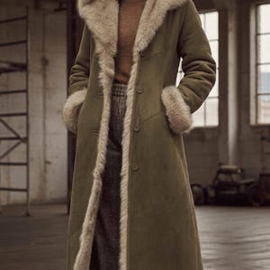 Może przedstawiać: Długi płaszcz w kolorze oliwkowym z kapturem i wykończeniem z imitacji futra. Płaszcz posiada zapięcie na guziki i kieszenie. Modelka ma na sobie brązowy golf i szare spodnie w kratę. Stylowa odzież wierzchnia.