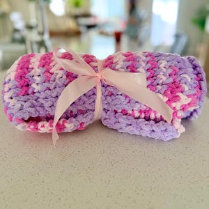 May include: A rolled-up, chunky knit blanket in shades of pink, purple, and white, tied with a light pink satin ribbon. The blanket appears soft and plush, suitable for a baby or as a decorative throw.