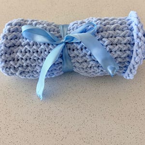 May include: A rolled-up, light blue, chunky knit blanket tied with a matching satin ribbon. The blanket has a soft, plush texture and is tightly rolled. The ribbon is tied in a bow, adding a decorative touch to the cozy item.