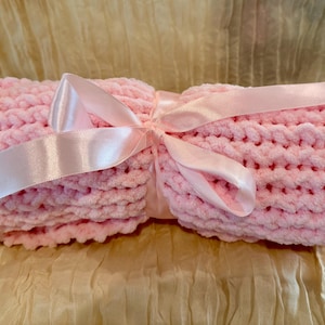 May include: A rolled-up, light pink, chunky knit blanket tied with a matching satin ribbon. The blanket is made of a soft, textured material, and the ribbon is tied in a bow. The blanket is a cozy item.