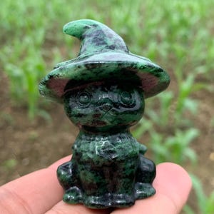 May include: A small carved figurine of a cat wearing a pointed hat, crafted from a dark green stone with black speckled inclusions. The cat has large, rounded eyes and a seated posture, showcasing the natural mottled texture of the polished mineral.