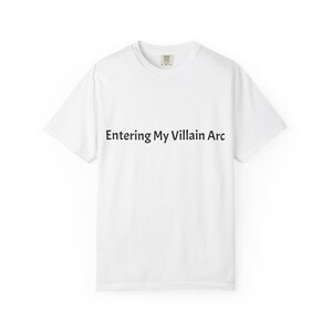 May include: A white t-shirt featuring the black text "Entering My Villain Arc" across the chest. The t-shirt has a crew neck and short sleeves. The material looks to be a soft cotton blend, suitable for casual wear.