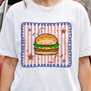 May include: A white short-sleeve t-shirt featuring a central graphic of a cheeseburger centred within a square frame. The background of the graphic consists of vertical red stripes, blue stars, and blue swirls, all enclosed by a blue scalloped border.