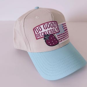 May include: A beige baseball cap with a light blue peak and top button. The cap has a patch with the text "DO GOOD RECKLESSLY", a strawberry, and an American flag design.