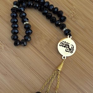 Może przedstawiać: Czarny różaniec z fasetowanymi koralikami ze złotym wisiorkiem z arabskim pismem. Koraliki są nawleczone na złoty łańcuszek i chwost. Koraliki spoczywają na drewnianej powierzchni.