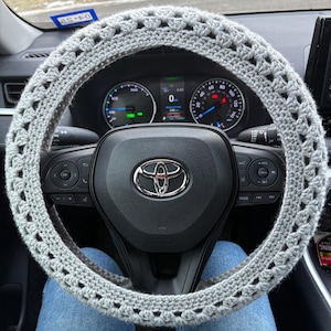 May include: A light gray, crocheted steering wheel cover. The cover is handmade with a repeating openwork pattern. The steering wheel is black with a silver Toyota emblem in the center. The car's dashboard and a person's blue jeans are visible.
