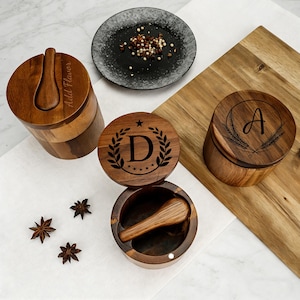 May include: Wooden spice jars with engraved lids, one with the letter "A" and wheat, another with "D" inside a laurel wreath, and a third with a spoon and the text "Add Flavor". A small black plate holds peppercorns. Star anise is scattered around.