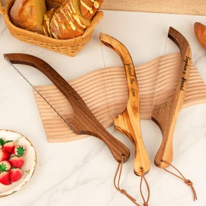 Puede incluir: Tres cortadores de pan de madera con mangos curvos y cuchillas de acero inoxidable. Uno es marrón oscuro con el texto "Best Ever Sliced Bread", otro es marrón claro con "Spread the Love" y el tercero es marrón claro con "Sophia's Bread".