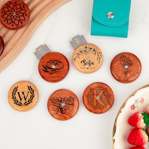 Könnte beinhalten: Eine Sammlung von Rasierklingenhaltern aus Holz mit eingravierten Designs. Die Halter weisen verschiedene Muster auf, darunter Blumenarrangements, Initialen und eine Biene. Ein Halter trägt den eingravierten Namen "Sofia". Ein türkisfarbenes Lederetui ist ebenfalls vorhanden.