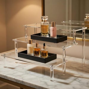 May include: A three-tiered clear acrylic display stand with black trays holding various perfume bottles. The bottles are different shapes and sizes, with gold and silver accents. The stand is on a marble surface.