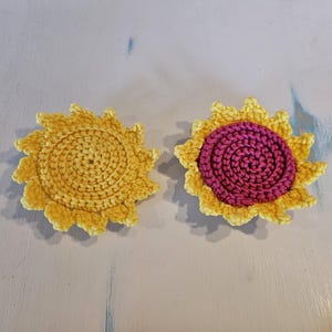 May include: Two handmade crochet sunflower coasters. One is solid yellow, the other has a pink centre. The coasters have a textured, three-dimensional appearance, with a spiral centre and petal-like edges. They are placed on a light blue and white surface.