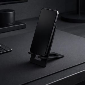 Könnte beinhalten: Ein schwarzes Smartphone wird auf einem schwarzen Telefonständer angezeigt. Der Ständer ist abgewinkelt und hält das Telefon aufrecht. Der Hintergrund umfasst einen schwarzen Schreibtisch, eine Tastatur und anderes dunkles Zubehör, was eine minimalistische Ästhetik schafft.
