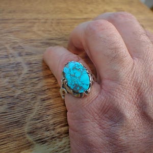 May include: A silver ring with a large, oval turquoise stone. The stone is a vibrant blue-green color with dark matrix patterns. The ring is worn on a finger, showcasing the intricate details of the setting.