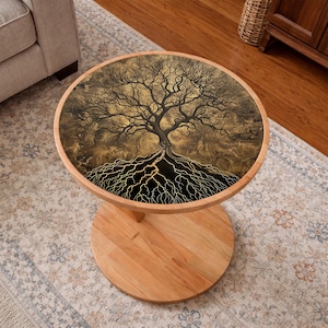 May include: A round wooden side table with a tree of life design on the tabletop. The tree is depicted in black and gold, with intricate branches and roots. The table has a light wood finish and a pedestal base.