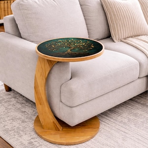 May include: A round side table with a wooden base and a dark teal top featuring a gold tree design. The table has a curved leg that allows it to slide under a sofa. The table is approximately 20 inches in diameter.
