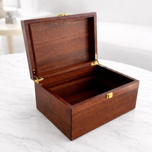 May include: A wooden box with a hinged lid, open to reveal the interior. The box is a rich, dark brown color with visible wood grain. Gold-colored hardware, including hinges and a clasp, adds a touch of elegance. The box is empty.