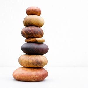 May include: A stack of seven smooth, rounded wooden stones in varying shades of brown and tan. The stones are balanced on top of each other, creating a tower against a white background. The wood grain is visible on some stones.