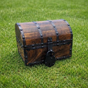 May include: A wooden treasure chest with a dark brown finish and black metal accents. The chest has a curved top, metal bands, and a black padlock. It is sitting on a bed of green grass.