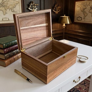 May include: An open wooden box with a hinged lid, showcasing a natural wood grain. The box features brass hardware and is set on a white surface, with a gold pen and a pocket watch nearby. The interior of the box is visible.