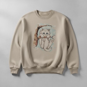 Könnte beinhalten: Beiges Sweatshirt mit einer skurrilen Illustration eines flauschigen, weißen Kätzchens, das sich an einem Ast festhält. Das Kunstwerk hat einen Vintage-Stil, handgezeichnet, mit sanften Farben und einem hellblauen Hintergrund.