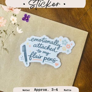 May include: A vinyl sticker with a white background featuring the dark blue text 'emotionally attached to my flair pens' alongside a blue pen illustration, small colorful flowers, and a heart. The sticker measures approximately 3-4 inches.