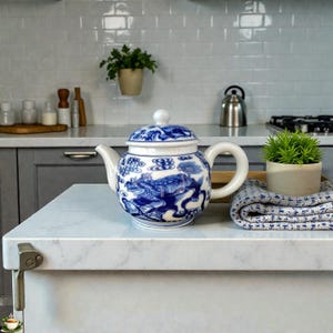 Op de afbeelding: Een wit en blauw keramieken theepot met een deksel, met een gedetailleerd drakenontwerp. De theepot heeft een gebogen handvat en tuit. De achtergrond omvat een keukenblad, een kleine plant en een opgevouwen handdoek.