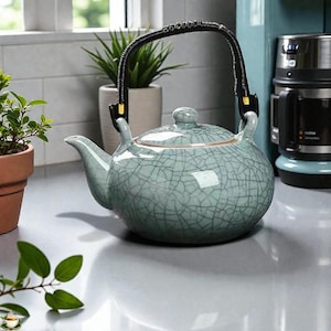 May include: A light blue ceramic teapot with a crackle glaze pattern. It has a black handle and a lid with a small knob. The teapot is on a countertop near a window and a coffee maker.