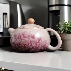 Puede incluir: Tetera de cerámica rosa claro con un gran diseño floral de peonías rosas. La tetera tiene un cuerpo redondeado, un asa curva y un pequeño pomo de madera en la tapa. Una pequeña planta en maceta está a la derecha.