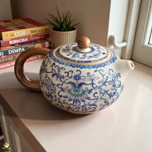 Könnte beinhalten: Eine weiße Keramik-Teekanne mit Holzgriff und Deckelknopf. Die Teekanne ist mit blauen Blumenmustern verziert. Der Griff hat eine hellbraune Farbe. Die Teekanne steht auf einer weißen Oberfläche.