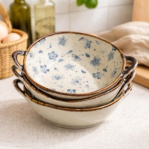 Puede incluir: Tres cuencos de cerámica apilados, con un diseño floral azul sobre un fondo crema. Los cuencos tienen bordes y asas marrones. Adecuados para servir alimentos.